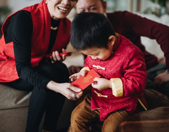 Cute little grandson dressed in red traditional Chinese costume receives red envelops (lai see) with both hands from grandparents joyfully in Chinese New Year